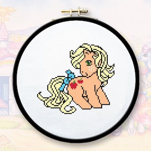 Puede incluir: Un aro de bordado de punto de cruz con una imagen pixelada de un pony marrón claro con una melena y cola rubias. El pony tiene un lazo azul y tres manzanas rojas en su flanco.