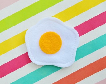 May include: A white felt egg with a yellow felt yolk, on a background of colorful stripes.