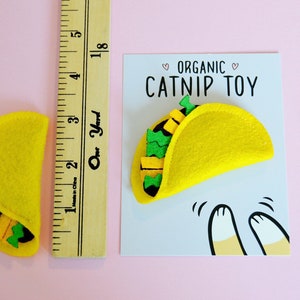 May include: A yellow felt catnip toy shaped like a taco with green and yellow felt filling. The toy is on a white card with the text "Organic Catnip Toy" in black.