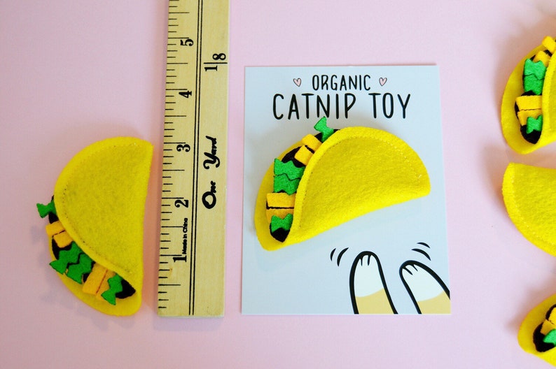 May include: A yellow felt catnip toy shaped like a taco with green and yellow felt filling. The toy is on a white card with the text "Organic Catnip Toy" in black.