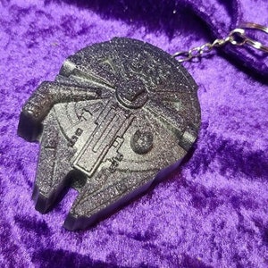 May include: A silver-colored Millennium Falcon keychain with a glittery finish. The keychain is attached to a silver chain.
