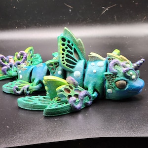 Puede incluir: Una figurita de criatura marina impresa en 3D con detalles en verde, azul y morado. La criatura tiene alas, cola y un cuerno. Est&aacute; hecha de pl&aacute;stico y tiene una superficie texturizada.