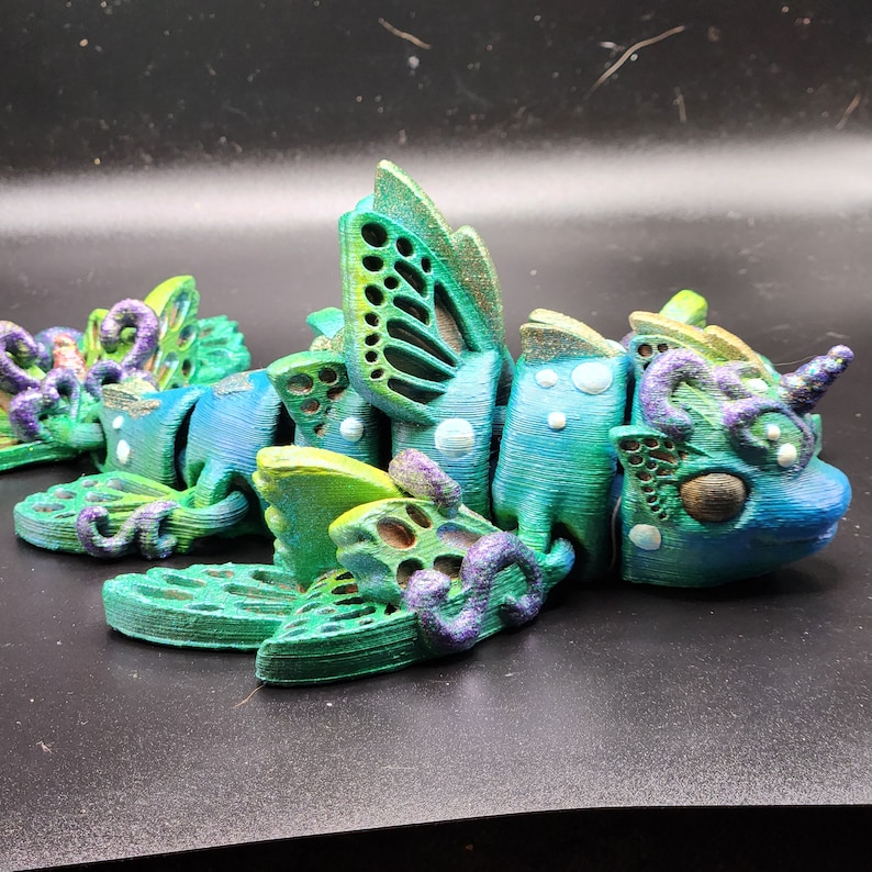 Puede incluir: Una figurita de criatura marina impresa en 3D con detalles en verde, azul y morado. La criatura tiene alas, cola y un cuerno. Est&aacute; hecha de pl&aacute;stico y tiene una superficie texturizada.