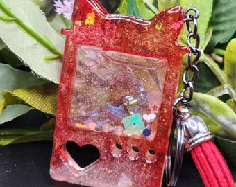 Peut inclure: Porte-clés en résine rouge pailletée en forme de console de jeu portable rétro. Le porte-clés a un centre transparent rempli de paillettes colorées et de petits charmes. Une découpe en forme de cœur et trois petits cercles sont visibles.