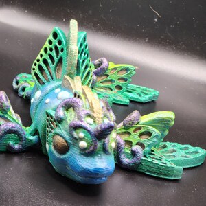 Puede incluir: Una figurita impresa en 3D de una criatura fant&aacute;stica con alas verde y azul iridiscentes, un cuerpo dorado y morado y detalles blancos. La criatura tiene una apariencia caprichosa y de hada.