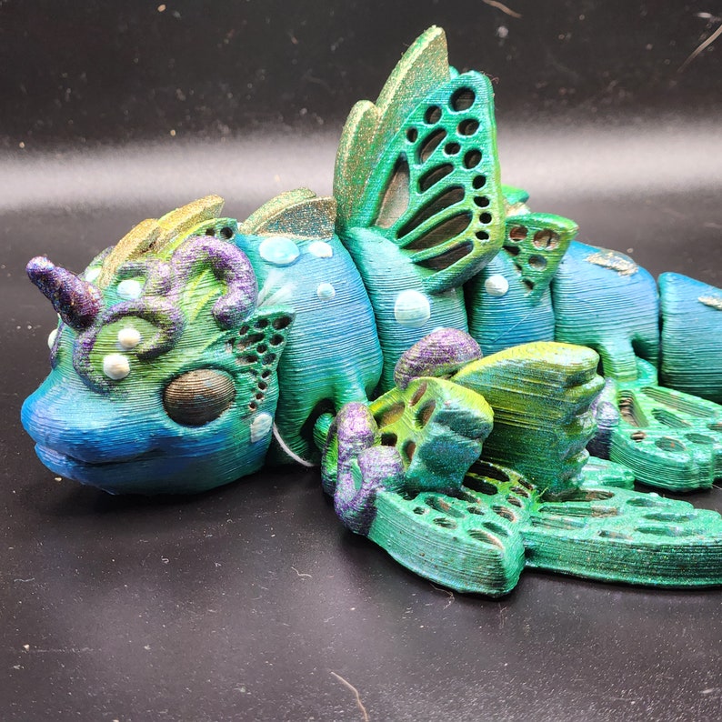 Puede incluir: Una figurita de drag&oacute;n impresa en 3D, azul, verde y morado con un dise&ntilde;o de ala de mariposa. El drag&oacute;n tiene un cuerno y una cara detallada.