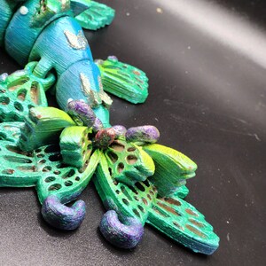 Puede incluir: Una figurita impresa en 3D de una criatura azul y verde con detalles intrincados. La criatura tiene alas y una cola que se asemejan a una mariposa. La figurita est&aacute; pintada con colores iridiscentes y tiene un acabado brillante.