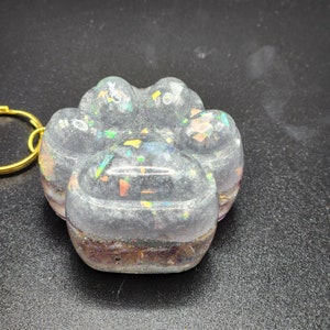 Puede incluir: Un llavero transparente con forma de huella de pata con purpurina iridiscente incrustada en la resina. El llavero está unido a un anillo de metal dorado.