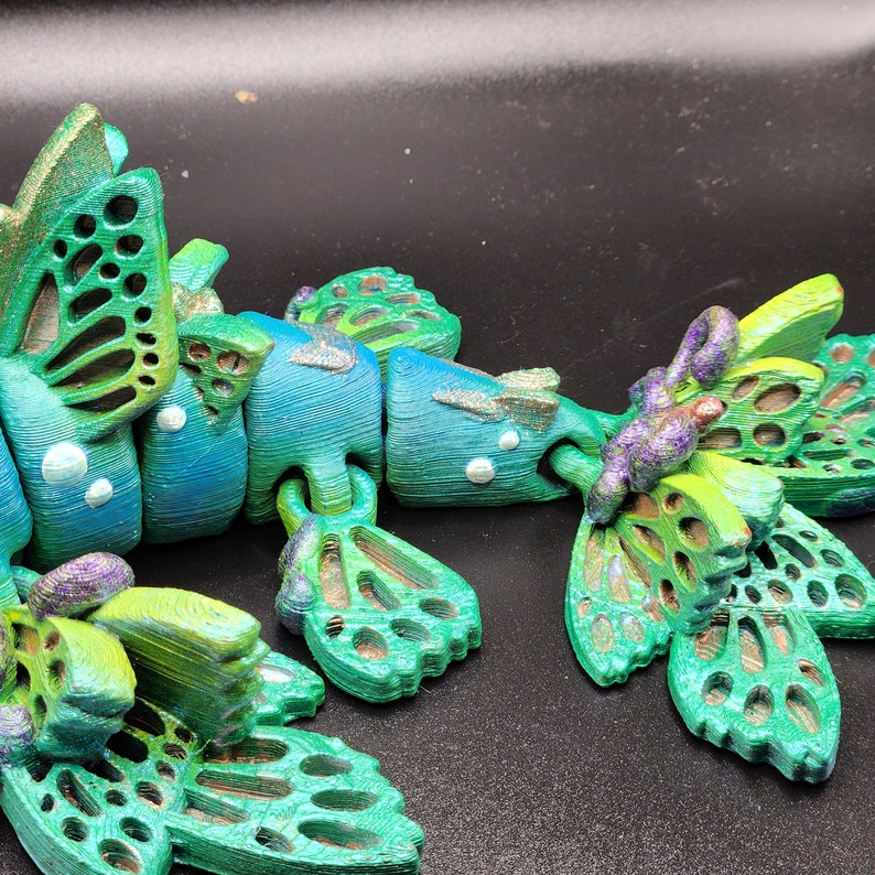 Puede incluir: Un juguete articulado verde y azul con un dise&ntilde;o de mariposa. El juguete est&aacute; hecho de pl&aacute;stico y tiene un cuerpo segmentado con m&uacute;ltiples alas.