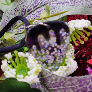 Puede incluir: Un colgante en forma de corazón con un fondo de resina transparente y purpurina incrustada. La purpurina es en tonos de rosa, morado y azul. El colgante está colgado de un cordón negro.