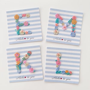 Può includere: Quattro biglietti quadrati a righe blu e bianche presentano lettere floreali ricamate: E, M, K e L. Ogni lettera è composta da fiori e foglie colorate. Il testo "Aloha to you" è stampato sotto ogni lettera.
