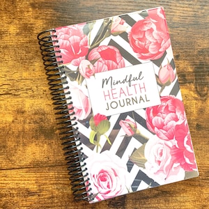 May include: A black and white spiral bound journal with a floral design featuring pink roses and peonies. The cover has the text "Mindful Health Journal" in black.