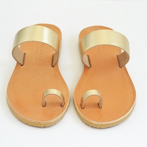 May include: A pair of gold leather toe ring sandals with a brown leather sole. The sandals have a simple design with a single strap across the top of the foot.