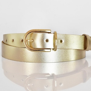 Op de afbeelding: Een gouden leren riem met een gouden metalen gesp. De riem is opgerold en de gesp is zichtbaar.