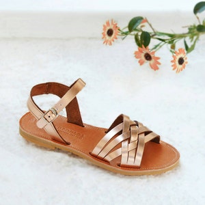 May include: Rose gold leather sandals with a woven strap design and ankle strap with buckle closure. The sandals have a brown leather footbed and a light brown sole. The sandals are displayed on a white surface with flowers in the background.