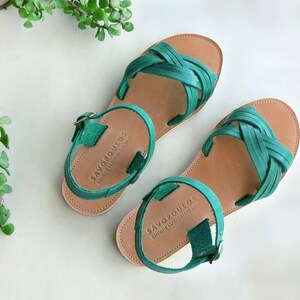 May include: A pair of green leather sandals with a woven strap design. The sandals have a brown leather sole and a buckle closure. The brand name "SAVOPOULOS" is printed on the straps.