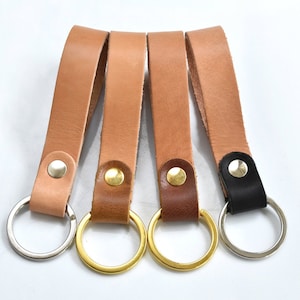 May include: Four leather keyrings in shades of brown and black. Each keyring has a leather strap attached to a metal ring. The metal rings are silver and gold-toned, and the straps are secured with metal rivets.