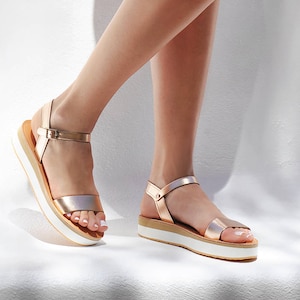 May include: A pair of gold platform sandals with a white sole. The sandals have a single strap across the foot and an ankle strap.