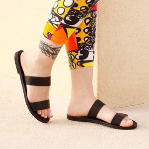 Puede incluir: Un par de sandalias de cuero negro con dos correas. Las sandalias son usadas por una persona que lleva leggings coloridos.