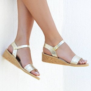 May include: A pair of gold wedge sandals with a cork platform and ankle straps. The sandals have a perforated design and are worn on a person's feet.