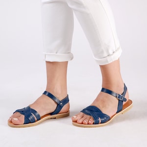 May include: A pair of blue leather sandals with a criss-cross strap design and a buckle closure. The sandals have a flat sole and are worn by a person with white pants.