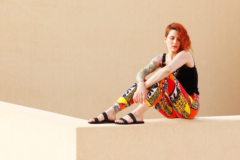 Puede incluir: Una mujer con el pelo rojo se sienta en una superficie de color beige claro con sandalias negras, un top negro sin mangas y pantalones estampados de colores.