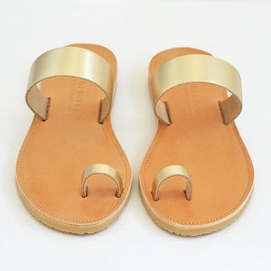 Peut inclure: Une paire de sandales en cuir or avec une lani&egrave;re de toe ring et une semelle en cuir marron. Les sandales ont une seule sangle sur le dessus du pied et une lani&egrave;re de toe ring.