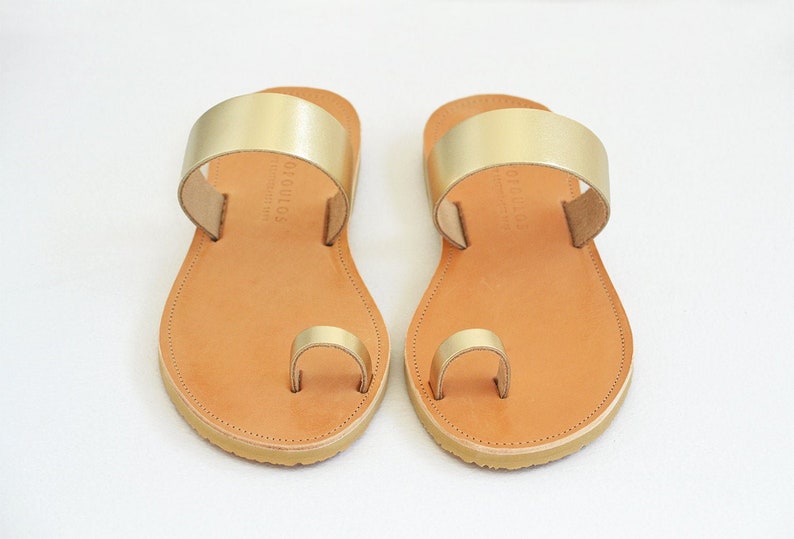 Peut inclure: Une paire de sandales en cuir or avec une lani&egrave;re de toe ring et une semelle en cuir marron. Les sandales ont une seule sangle sur le dessus du pied et une lani&egrave;re de toe ring.