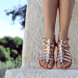 Puede incluir: Un par de sandalias gladiadoras doradas con m&uacute;ltiples correas y hebillas. Las sandalias son usadas por una persona de pie sobre una superficie de piedra.
