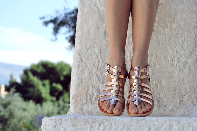 Puede incluir: Un par de sandalias gladiadoras doradas con m&uacute;ltiples correas y hebillas. Las sandalias son usadas por una persona de pie sobre una superficie de piedra.