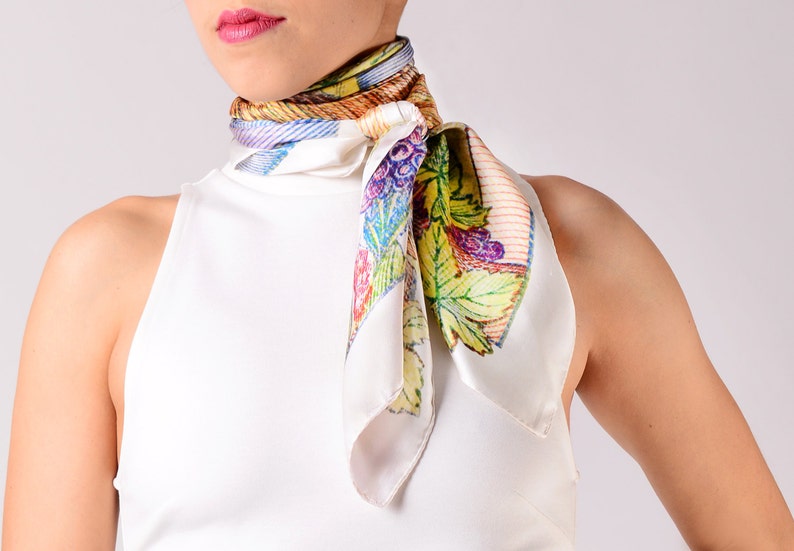 May include: A white silk scarf featuring a colourful grape and leaf design. The scarf is tied around the neck, displaying a vibrant pattern of purple grapes, green leaves, and a striped border. The scarf adds a pop of colour.