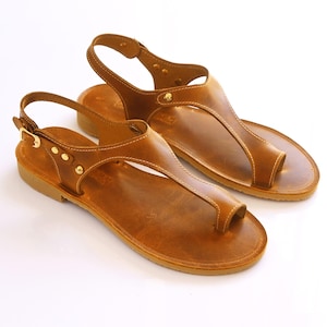 May include: A pair of tan leather sandals featuring a thong-style toe post, an adjustable ankle strap with a gold-toned metal buckle, and decorative gold-toned rivets along the side straps. The sandals have a flat sole and visible stitching.