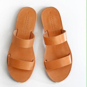Puede incluir: Un par de sandalias de cuero marr&oacute;n con dos correas. Las sandalias tienen el texto "SAYOPOULOS" en las correas.