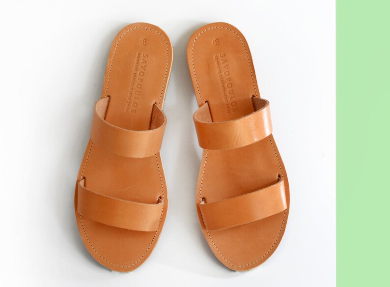 Puede incluir: Un par de sandalias de cuero marr&oacute;n con dos correas. Las sandalias tienen el texto "SAYOPOULOS" en las correas.