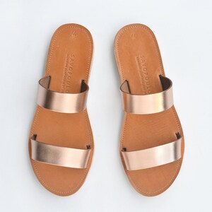 Puede incluir: Un par de sandalias de cuero color oro rosa con dos tiras. Las sandalias tienen una suela de cuero marr&oacute;n y est&aacute;n hechas por Savopoulo.