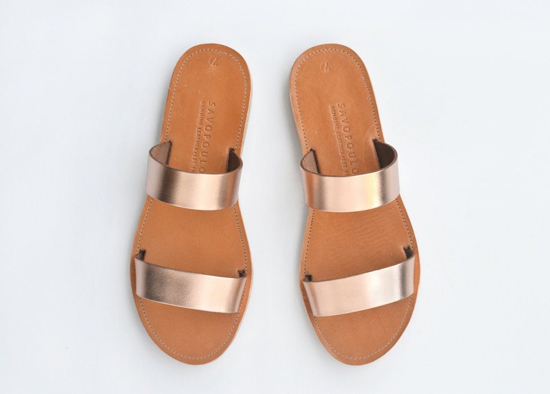 Puede incluir: Un par de sandalias de cuero color oro rosa con dos tiras. Las sandalias tienen una suela de cuero marr&oacute;n y est&aacute;n hechas por Savopoulo.