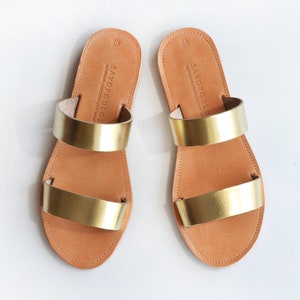 Puede incluir: Un par de sandalias de cuero dorado con dos tiras. Las sandalias est&aacute;n hechas por Savopoulos.