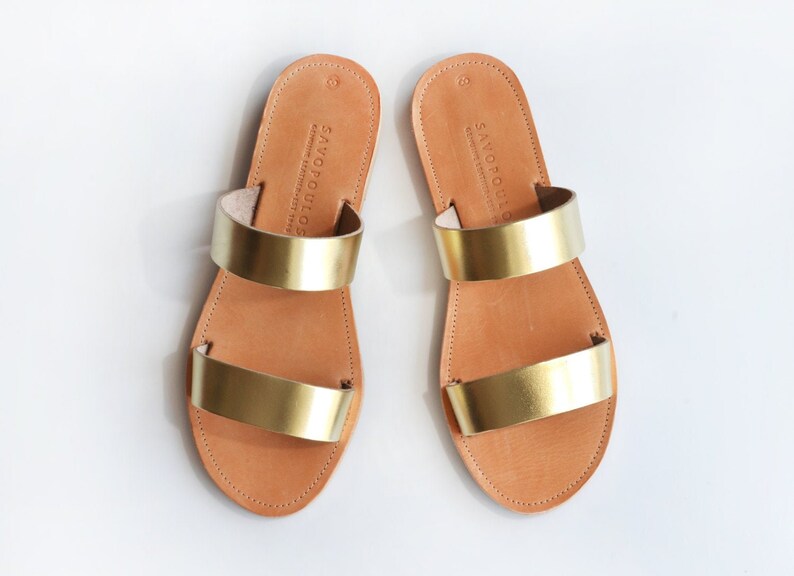 Puede incluir: Un par de sandalias de cuero dorado con dos tiras. Las sandalias est&aacute;n hechas por Savopoulos.