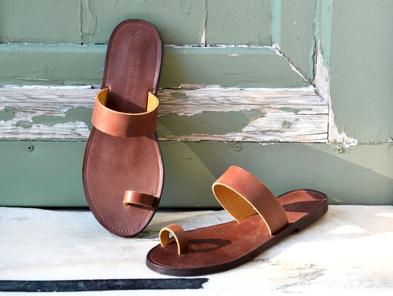 Peut inclure: Une paire de sandales en cuir marron avec une boucle d'orteil et une seule sangle sur le pied. Les sandales ont une doublure en cuir marron clair.