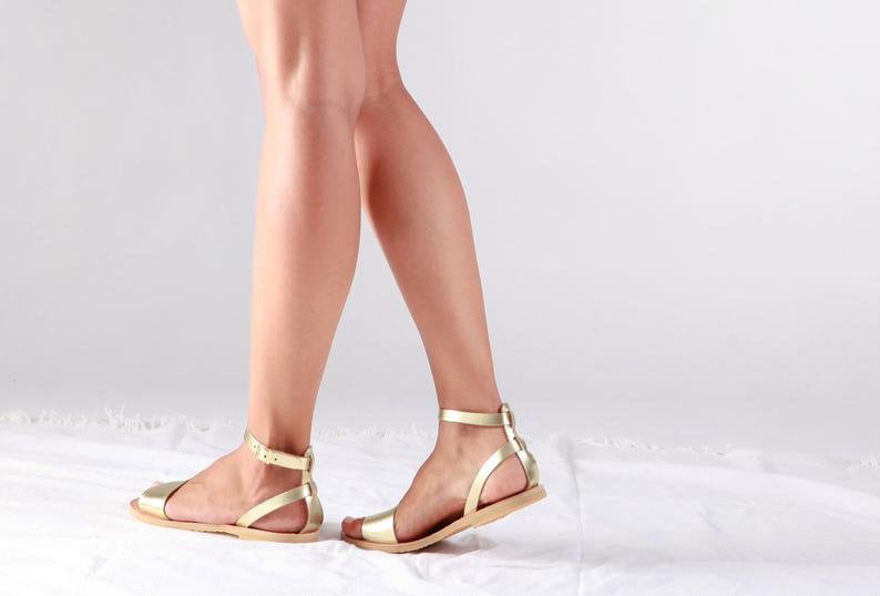 May include: A pair of gold leather sandals with ankle straps. The sandals have a flat sole and are made of a soft, supple leather.