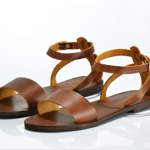 May include: A pair of brown leather sandals with ankle straps and buckles. The sandals have a flat sole and a simple design.