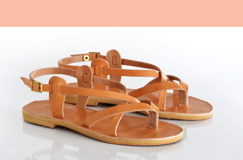 May include: A pair of tan leather sandals with criss-cross straps and a buckle closure. The sandals have a flat sole and a toe loop.