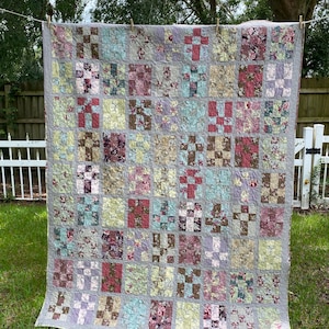 Può includere: Una trapunta patchwork rettangolare caratterizzata da una griglia di blocchi alternati. La trapunta è composta da vari quadrati di tessuto floreale e fantasia in tonalità di rosa tenue, verde salvia, marrone e crema, uniti da cuciture visibili.