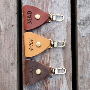Könnte beinhalten: Drei Leder-Schlüsselanhänger mit Metallschließen. Die Schlüsselanhänger sind dreieckig und haben die Worte "Mad", "Rock" und "Luck" eingraviert.