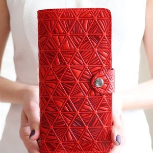 Puede incluir: Una vibrante cartera roja con un patrón geométrico de triángulos. La cartera es rectangular y tiene un cierre de botón a presión. El diseño está en relieve, creando una superficie texturizada. La cartera se sostiene frente a un fondo blanco.