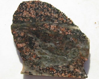 May include: A close-up of a rough, irregularly shaped rock with a mix of black, green, and pink colors. The rock has a layered appearance with white veins running through it.