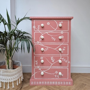 May include: A pink chest of drawers with five drawers and white floral vine detailing. The chest has white round knobs and decorative trim. A potted plant in a woven basket is to the left of the chest.