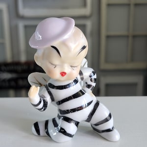 May include: A vintage ceramic figurine of a kneeling cherub with closed eyes and a red mouth. The cherub wears a light purple hat and a black and white striped outfit. The figurine has small, detailed wings.