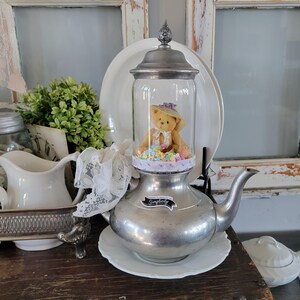May include: A silver teapot with a glass dome lid containing a teddy bear wearing a hat and surrounded by pastel-colored candy. The teapot has a label that reads "Embrace Simplicity".