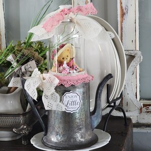 May include: A vintage silver teapot with a pink and white lace trim, a glass cloche with a teddy bear inside, and a sign that reads "Enjoy the Little Things".